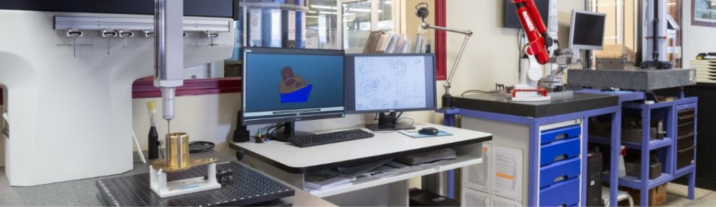 plan large sur le bureau d'études, avec deux écrans d'ordi montrant des pièces en 3D.